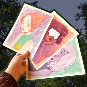 Può includere: Mano che tiene tre stampe artistiche ad acquerello con personaggi di una serie animata. Ogni stampa mostra un personaggio diverso con caratteristiche e palette di colori uniche, su sfondi verdi e viola.