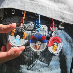 Könnte beinhalten: Drei kreisförmige Acryl-Schlüsselanhänger mit Miniatur-Charakterillustrationen in stilisierten Ball-Designs. Jeder Schlüsselanhänger hängt an einem farbigen Kunststoffclip, der an einem schwarzen Gürtel befestigt ist. Die Charaktere umfassen eine hellblaue, eine beige und eine gelbe Kreatur.