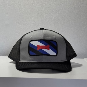 Puede incluir: Gorra de camionero gris y negra con un parche rectangular con un hueso rojo sobre rayas azules, negras y blancas. La gorra tiene una visera negra y una parte trasera de malla.