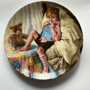 May include: A decorative plate depicting a child in a pink shirt and brown shorts, holding a book titled "Bear Stories." A teddy bear sits on a colourful quilt. The plate has a gold rim and is signed by the artist.