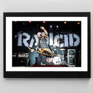 Puede incluir: Fotografía enmarcada de una actuación musical en directo. Un músico con barba toca una guitarra eléctrica dorada en el escenario. El nombre de la banda, "RANCID", se muestra en letras blancas grandes en el telón de fondo.