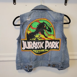 Può includere: Un giubbotto di jeans blu con un effetto consumato, con un logo Jurassic Park dipinto a mano sul retro. Il logo è giallo, rosso, verde e nero con una silhouette di dinosauro.