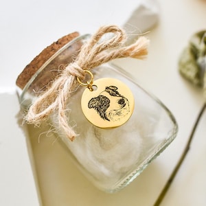 Peut inclure: Un bocal en verre avec un couvercle en liège et un charme en métal doré attaché. Le charme est un disque rond avec une image en noir et blanc d'un visage de chien gravée dessus. Le bocal est rempli de coton blanc.