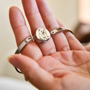 Könnte beinhalten: Ein silberfarbenes Metallarmband mit einem runden Anhänger, der eine detaillierte schwarz-weiße Illustration eines flauschigen Hundes zeigt. Der Anhänger ist zwischen zwei polierten Metallbändern zentriert, die mit kleinen Ringen verbunden sind.