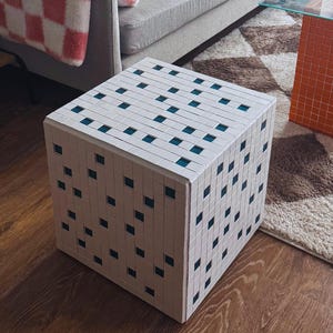 May include: A white cube-shaped ottoman with a pattern of small teal squares. The ottoman sits on a wooden floor in front of a gray sofa. A glass-top coffee table with orange tile legs is in the background.