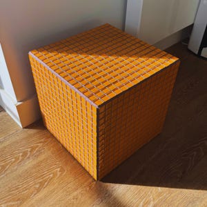 May include: A square orange mosaic tile cube, likely a stool or side table. The surface is covered in small, square orange tiles, with a light purple edge. The cube sits on a wooden floor, with sunlight casting shadows.