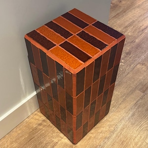 Puede incluir: Una mesa pedestal decorativa y rectangular con un patrón geométrico. La parte superior presenta filas alternas de baldosas naranjas y marrones oscuras. Los lados están cubiertos con un patrón repetido de baldosas marrones oscuras y rojizas. La mesa mide unos 61 cm de alto.