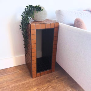 Può includere: Un tavolino rettangolare rivestito di piastrelle quadrate color arancio bruciato lucido, con una cavità interna rivestita di piastrelle nere. Un oggetto decorativo sferico di colore bianco sporco e dalla superficie ruvida poggia sul piano.