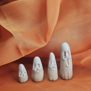 May include: Four small, off-white ghost figurines with sparkling accents on an orange background. The ghosts vary in size, with a simple, rounded design. The background is a textured orange fabric.