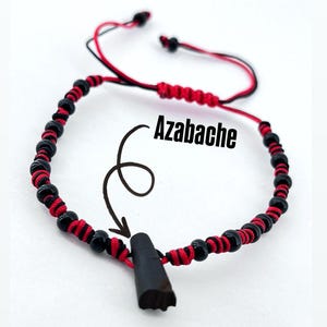 Puede incluir: Una pulsera de cuentas negras y rojas con un amuleto Azabache negro. La pulsera presenta cuentas negras y cordón rojo, con la palabra "Azabache" en la imagen. El amuleto tiene forma oscura y alargada.