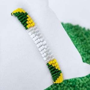 Może przedstawiać: Bransoletka z zielonych, żółtych i białych koralików. Bransoletka wykonana jest z małych, okrągłych koralików i ma prosty, geometryczny wzór.