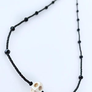 May include: A black beaded necklace with a white skull charm. The necklace is made of small black beads and has a single white skull charm in the center.