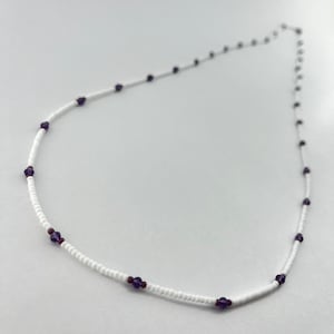 Puede incluir: Un delicado collar de cuentas blancas con pequeñas cuentas de color púrpura oscuro espaciadas uniformemente a lo largo del hilo. El collar se muestra sobre un fondo blanco liso, destacando su simple elegancia.