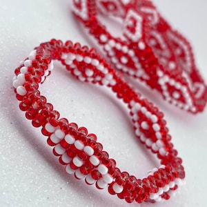 May include: Red and white beaded bracelet with a geometric pattern. The bracelet is made of small, round beads that are strung together in a repeating pattern.