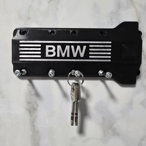 Könnte beinhalten: Schwarzer Schlüsselhalter in Form eines BMW-Motorblocks mit dem Schriftzug "BMW" in silbernen Lettern. Der Schlüsselhalter hat fünf Haken zum Aufhängen von Schlüsseln.