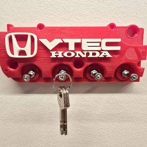 Puede incluir: Llavero rojo con los logotipos de Honda y VTEC en blanco. El llavero, con forma de componente de motor, tiene varios ganchos para llaves con herrajes plateados. Un juego de llaves cuelga de uno de los ganchos.