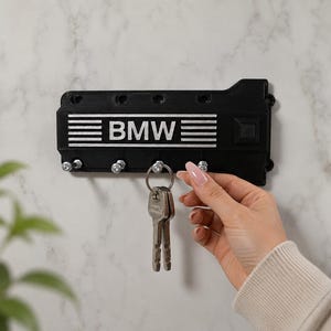 Può includere: Portachiavi nero con il logo BMW e strisce orizzontali bianche. Il portachiavi è montato a parete e ha ganci per le chiavi. Una mano tiene un mazzo di chiavi attaccate al portachiavi.