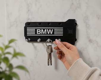 Op de afbeelding: Zwarte sleutelhouder met het BMW-logo en witte horizontale strepen. De sleutelhouder is aan een muur bevestigd en heeft haken voor sleutels. Een hand houdt een set sleutels vast die aan de sleutelhouder zijn bevestigd.