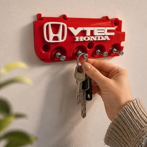Könnte beinhalten: Roter Honda VTEC Schlüsselhalter an einer weißen Wand. Der Schlüsselhalter hat das Honda-Logo und den VTEC-Text in Weiß. Ein Schlüsselbund wird in der Nähe des Schlüsselhalters gehalten.