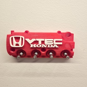 Puede incluir: Un llavero rojo con forma de componente de motor de coche, con el logotipo de Honda y el texto "VTEC HONDA" en blanco. Tiene cuatro ganchos plateados para llaves, diseñados para montaje en pared.