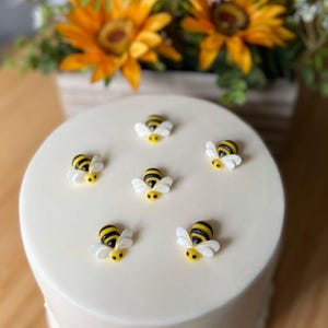 May include: A round, white cake adorned with six bee decorations. The bees feature black and yellow striped bodies and white wings. A bouquet of sunflowers and daisies is visible in the background.