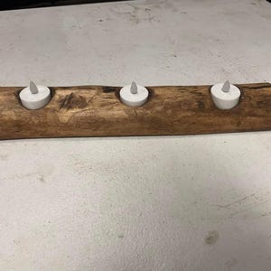 Puede incluir: Un portavelas rústico de madera con tres velas LED blancas. El soporte está hecho de una sección de un tronco, teñido de un color marrón cálido. Las velas están espaciadas uniformemente a lo largo de la madera.