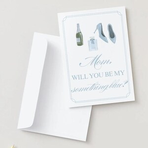 Puede incluir: Una tarjeta blanca con borde azul y texto "Mom, WILL YOU BE MY something blue?" La tarjeta presenta ilustraciones de acuarela de una botella de champán, perfume y tacones azules. Una sobre blanco abierto está al lado de la tarjeta.