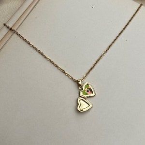 K&ouml;nnte beinhalten: Eine goldfarbene herzf&ouml;rmige Medaillon-Halskette. Das Medaillon ist ge&ouml;ffnet und zeigt ein kleines Bild. Die Halskette hat eine zarte Kette. Das Medaillon liegt auf einer wei&szlig;en Oberfl&auml;che.