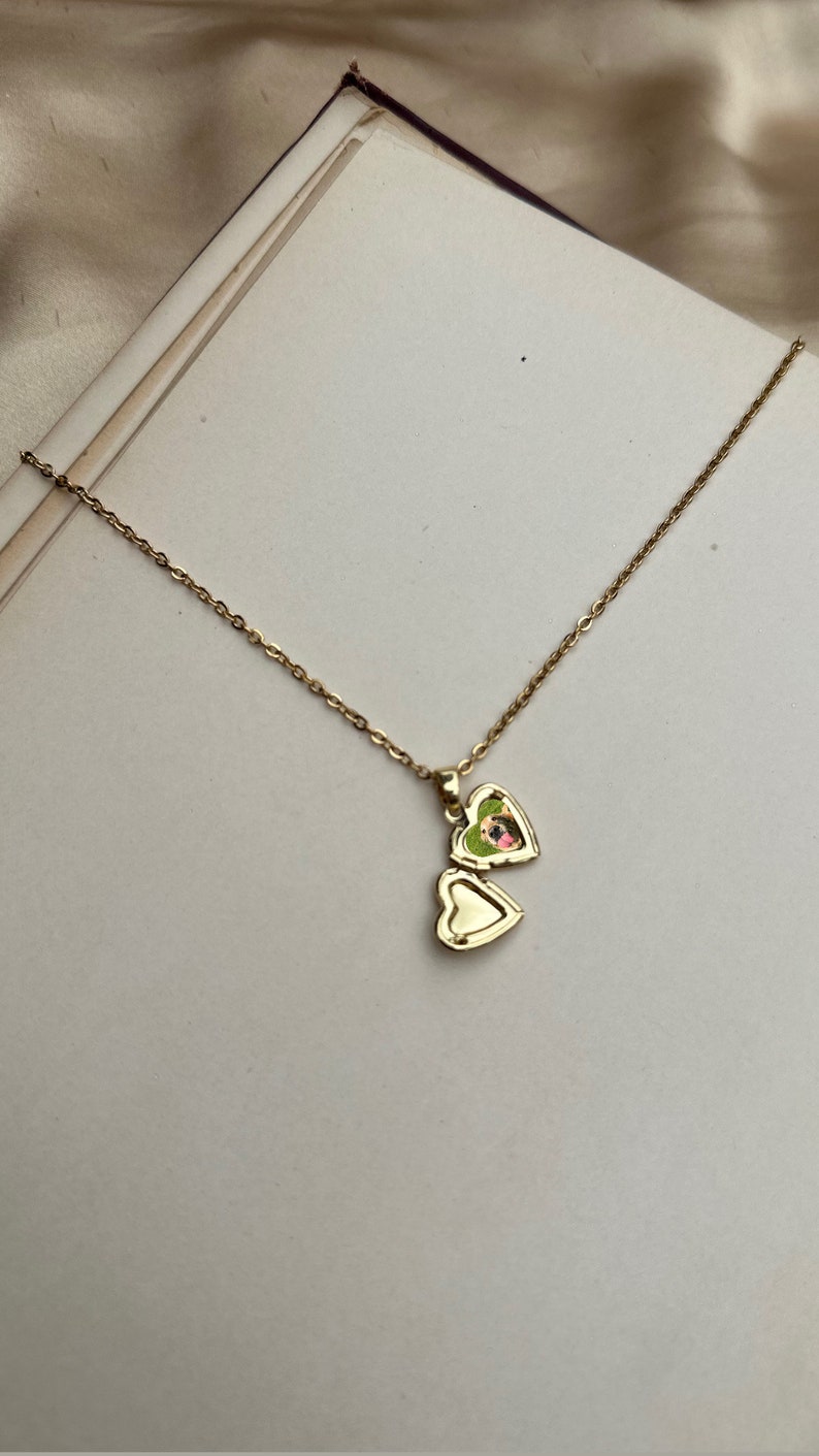 K&ouml;nnte beinhalten: Eine goldfarbene herzf&ouml;rmige Medaillon-Halskette. Das Medaillon ist ge&ouml;ffnet und zeigt ein kleines Bild. Die Halskette hat eine zarte Kette. Das Medaillon liegt auf einer wei&szlig;en Oberfl&auml;che.