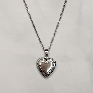 May include: A silver-colored heart-shaped locket pendant necklace. The locket has an ornate, decorative border and hangs from a delicate chain. The necklace is displayed against a light background.