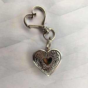 May include: A silver-colored heart-shaped locket with a decorative floral design, attached to a heart-shaped clasp. The locket is designed to hold a small photo or keepsake. The clasp is attached to a key ring.