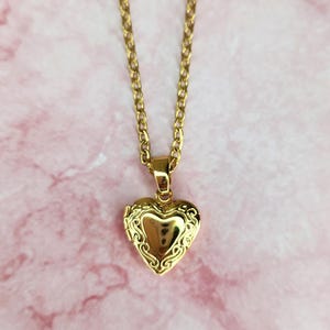 K&ouml;nnte beinhalten: Eine goldfarbene herzf&ouml;rmige Medaillon-Halskette. Das Medaillon hat ein verziertes Design an den R&auml;ndern und h&auml;ngt an einer zarten Kette. Der Hintergrund ist ein rosa-wei&szlig;es Marmormuster.