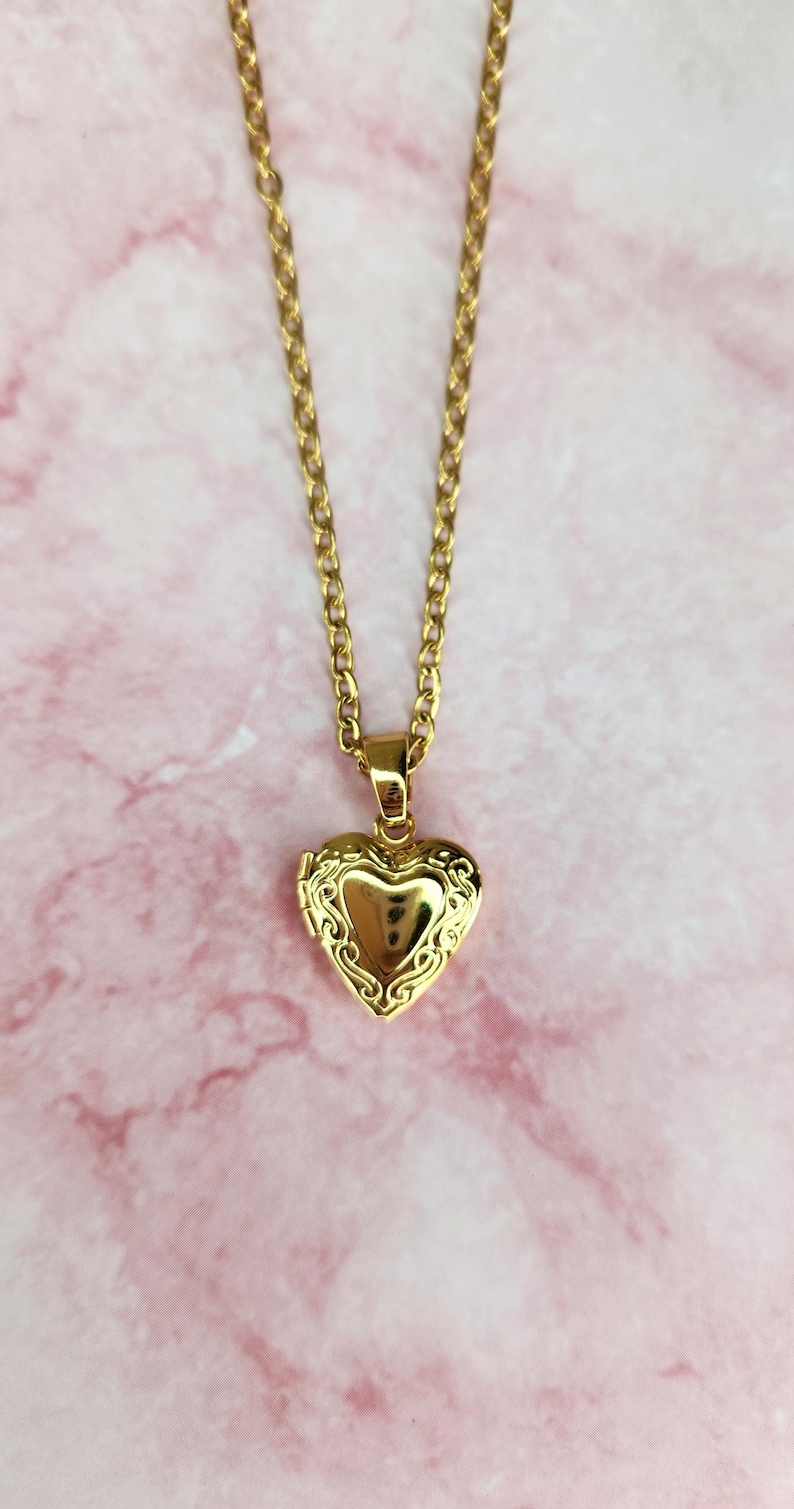 K&ouml;nnte beinhalten: Eine goldfarbene herzf&ouml;rmige Medaillon-Halskette. Das Medaillon hat ein verziertes Design an den R&auml;ndern und h&auml;ngt an einer zarten Kette. Der Hintergrund ist ein rosa-wei&szlig;es Marmormuster.