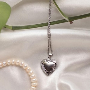 May include: A silver-toned heart-shaped locket pendant suspended from a delicate matching link chain. The locket features a smooth, polished center framed by an intricate, raised scrollwork border. The necklace is presented alongside a strand of white pearls.