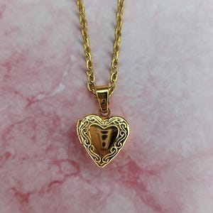 K&ouml;nnte beinhalten: Halskette mit herzf&ouml;rmigem, goldfarbenem Medaillon. Das Medaillon hat ein verziertes Filigranmuster an den R&auml;ndern und h&auml;ngt an einer passenden Kette. Der Hintergrund ist ein rosa Marmormuster.