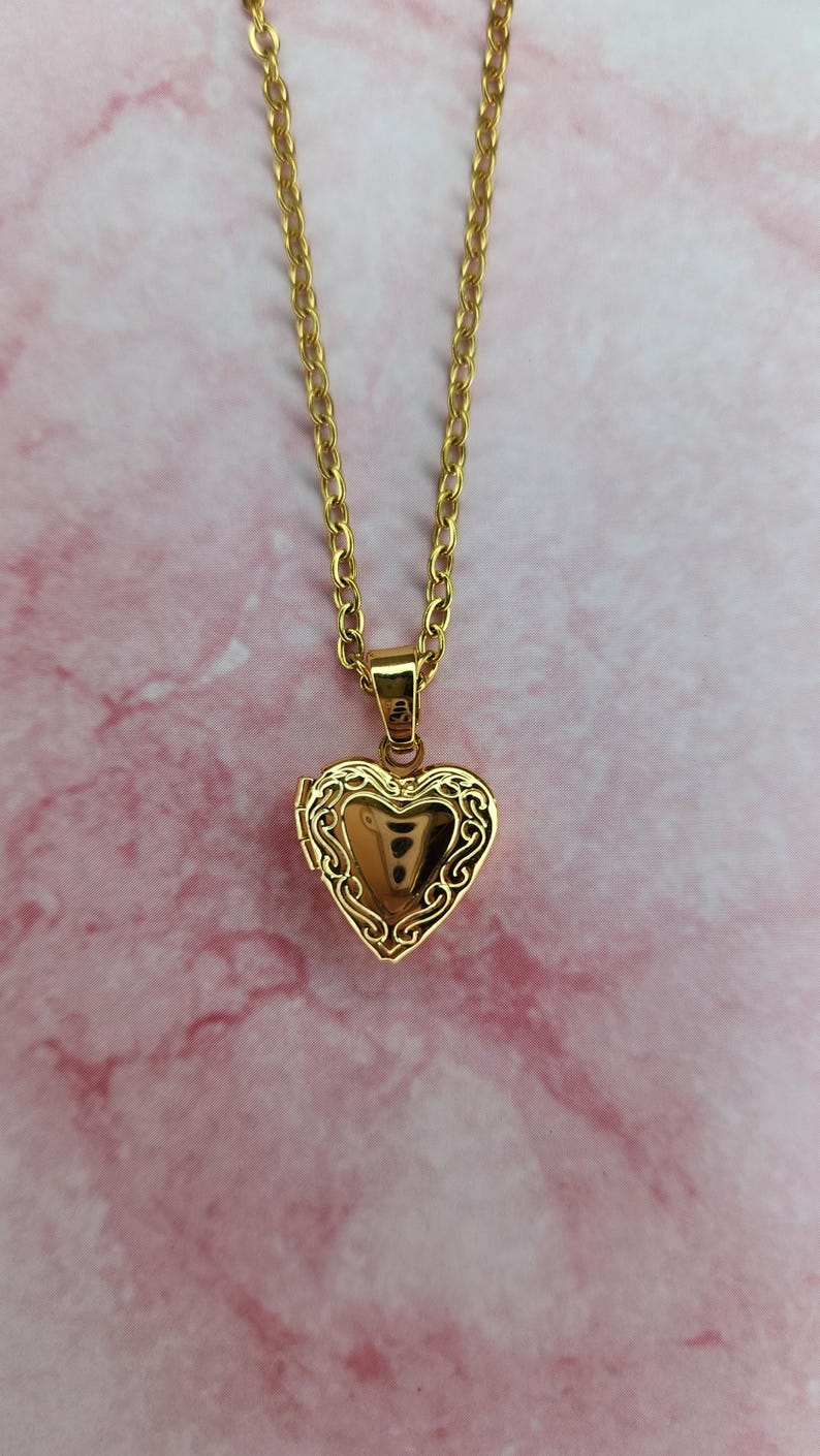K&ouml;nnte beinhalten: Halskette mit herzf&ouml;rmigem, goldfarbenem Medaillon. Das Medaillon hat ein verziertes Filigranmuster an den R&auml;ndern und h&auml;ngt an einer passenden Kette. Der Hintergrund ist ein rosa Marmormuster.