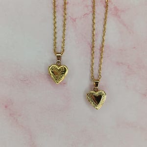 K&ouml;nnte beinhalten: Zwei goldfarbene herzf&ouml;rmige Medaillon-Halsketten mit filigranen Ketten. Die Medaillons haben aufwendige Blumendesigns und lassen sich &ouml;ffnen, um Platz f&uuml;r ein kleines Foto oder Andenken zu bieten. Der Hintergrund ist ein rosa-wei&szlig;es Marmormuster.