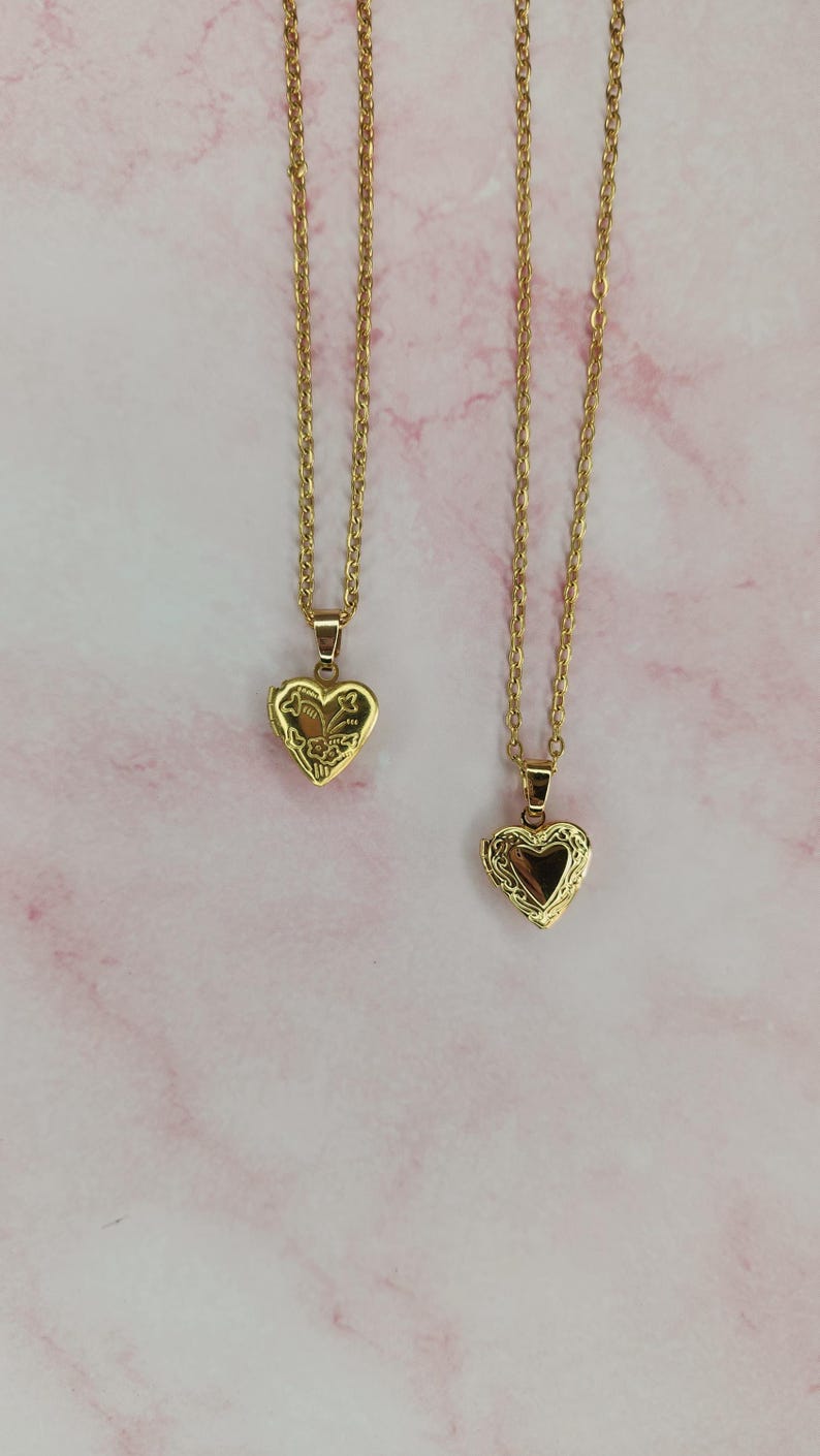 K&ouml;nnte beinhalten: Zwei goldfarbene herzf&ouml;rmige Medaillon-Halsketten mit filigranen Ketten. Die Medaillons haben aufwendige Blumendesigns und lassen sich &ouml;ffnen, um Platz f&uuml;r ein kleines Foto oder Andenken zu bieten. Der Hintergrund ist ein rosa-wei&szlig;es Marmormuster.