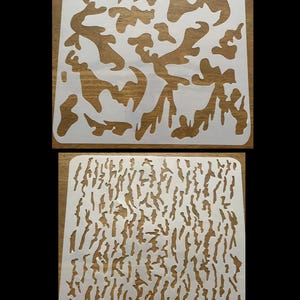 May include: Two square white plastic stencils featuring cut-out patterns. The top stencil displays large, irregular organic shapes. The bottom stencil features a dense, vertical pattern of thin, elongated, bark-like textured cut-outs.