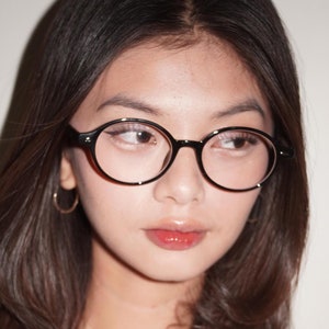 Puede incluir: Una joven con el pelo largo y castaño oscuro que lleva gafas redondas con montura negra. Lleva un jersey blanco con un botón de perla.