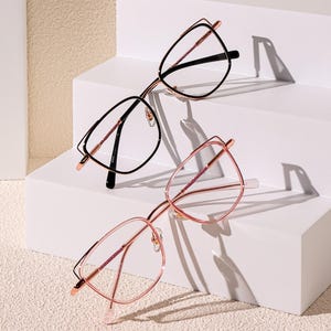 Op de afbeelding: Twee brillen met een cat-eye montuur. Het ene paar heeft een zwart montuur en heldere glazen. Het andere paar heeft een roségouden montuur en heldere glazen. De brillen zijn gerangschikt op een wit, meerlagig display.