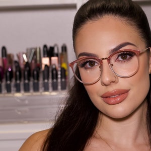May include: A woman with long dark hair wearing pink-rimmed glasses with clear lenses. She is wearing light makeup and has a neutral expression.