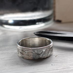 May include: A silver-toned metal ring featuring a band with an engraved pattern of curved lines and foliage motifs. The ring has a polished finish and a consistent width, with a detailed texture visible across the outer surface of the band.