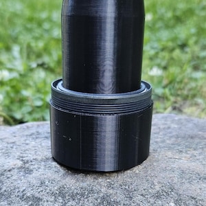 Puede incluir: Se muestra un objeto cilíndrico negro, posiblemente un contenedor o parte de un dispositivo. Tiene una base roscada y una sección superior lisa y más alta. El objeto está hecho de un material oscuro y sólido y se encuentra sobre una superficie de piedra gris, con follaje verde en el fondo.