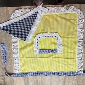 May include: A yellow and white striped apron with a matching navy blue and white checkered bandana. The apron has white lace trim and a pocket with blue trim. The apron ties are navy blue and white checkered.