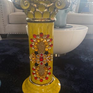 Puede incluir: Una columna decorativa amarilla con detalles dorados y plateados ornamentados. La columna presenta un patrón de piedras preciosas rojas, doradas y negras. La base y la parte superior de la columna también son amarillas con adornos dorados. Un artículo de decoración del hogar.