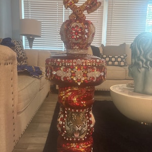 May include: A red and gold decorative stand with a tiger design and a crown on top. The stand is decorated with rhinestones and beads.
