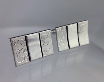 Könnte beinhalten: Ein Paar silberne Manschettenknöpfe mit rechteckigem Design. Jeder Manschettenknopf hat drei Abschnitte, wobei die äußeren Abschnitte ein geätztes, wirbelndes Blumenmuster aufweisen. Die Manschettenknöpfe werden auf einer reflektierenden Oberfläche präsentiert.