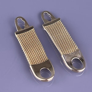 May include: Two gold-coloured zipper pulls with a mesh design. Each pull has a rectangular shape with a loop at the top and a rounded opening at the bottom. The pulls are set against a purple background.