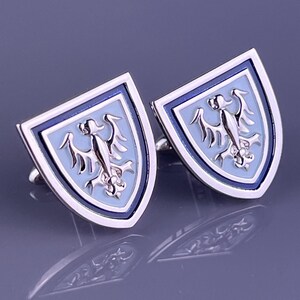 Puede incluir: Un par de gemelos de metal plateado con forma de escudos heráldicos. Cada escudo presenta un fondo de esmalte azul claro con un emblema de águila de plata pulida en relieve en el centro, enmarcado por un borde de esmalte azul oscuro.